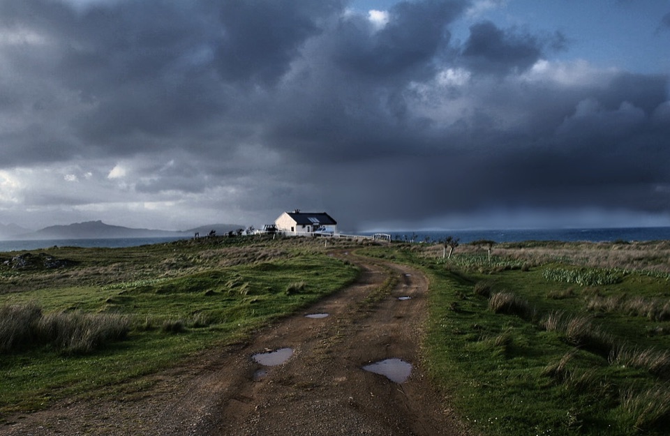 A white house standing alone on open moorland under a dark sky in the Highlands
